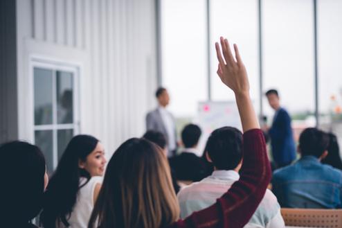 Bild zeigt eine Versammlung von Menschen in der eine Person sich meldet.